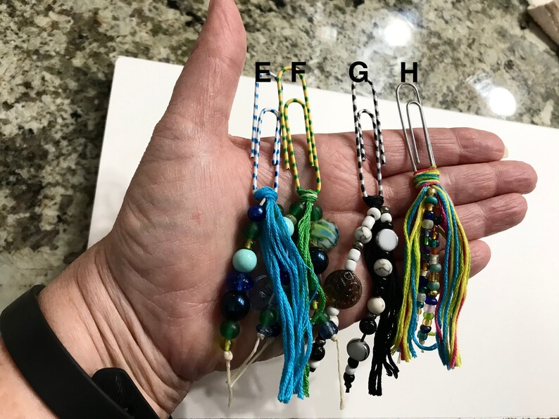 May include: Four colorful beaded and tassel bookmarks made with paper clips. Each bookmark has a different color scheme and bead combination. The bookmarks are held in a hand.
