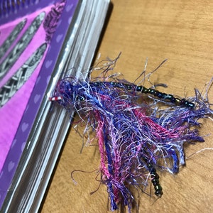 May include: A purple spiral bound notebook with a pink and white tassel bookmark. The tassel is made of frayed fabric and has small black beads.