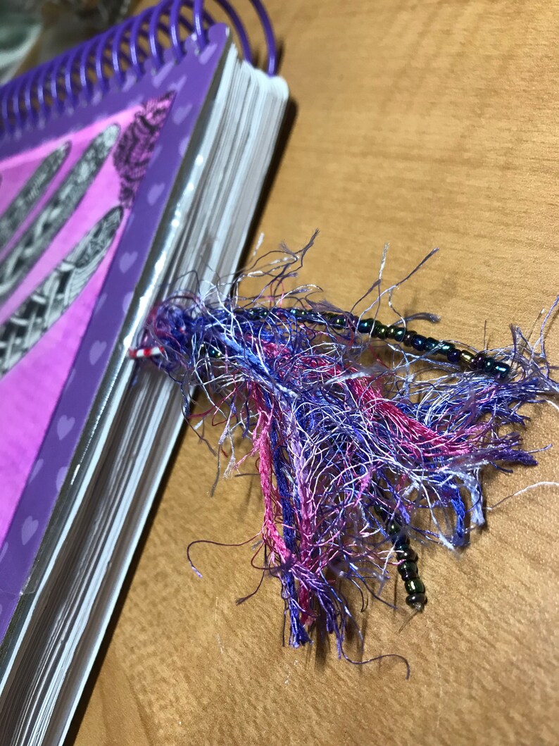 May include: A purple spiral bound notebook with a pink and white tassel bookmark. The tassel is made of frayed fabric and has small black beads.