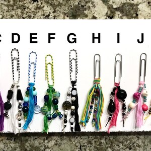 May include: A set of 12 colorful paper clip charms with beaded tassels. The charms are labeled A through L.
