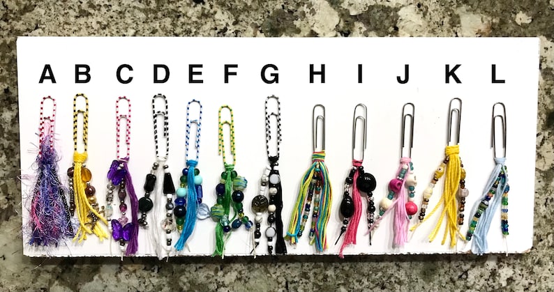May include: A set of 12 colorful paper clip charms with beaded tassels. The charms are labeled A through L.
