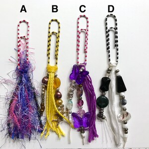 May include: Four colorful paper clips with different charms and beads hanging from them. The charms include purple butterflies, white tassels, and black and green beads.