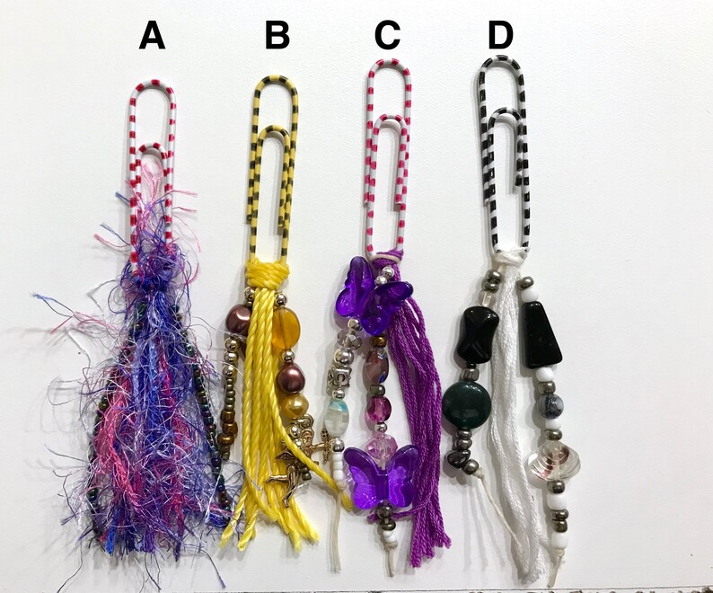 May include: Four colorful paper clips with different charms and beads hanging from them. The charms include purple butterflies, white tassels, and black and green beads.