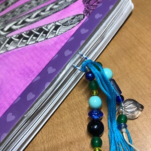 May include: A purple notebook with a spiral binding and a beaded tassel bookmark. The notebook has a heart pattern on the cover and a black and white drawing of feathers.