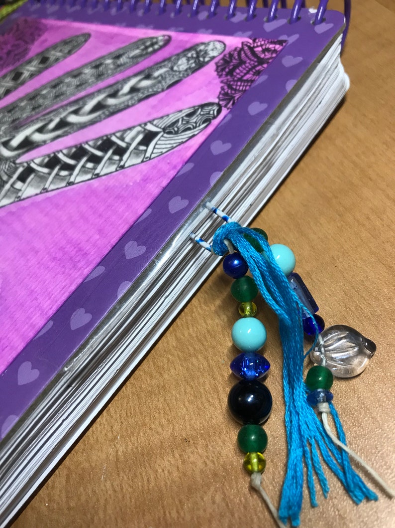 May include: A purple notebook with a spiral binding and a beaded tassel bookmark. The notebook has a heart pattern on the cover and a black and white drawing of feathers.