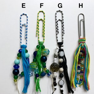 May include: Four colorful paper clips with beaded and tassel embellishments. The paper clips are decorated with stripes of blue and white, yellow and green, black and white, and yellow, blue, green, and pink.