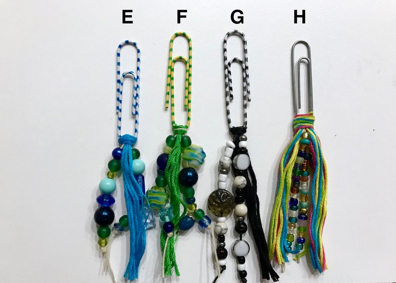 May include: Four colorful paper clips with beaded and tassel embellishments. The paper clips are decorated with stripes of blue and white, yellow and green, black and white, and yellow, blue, green, and pink.