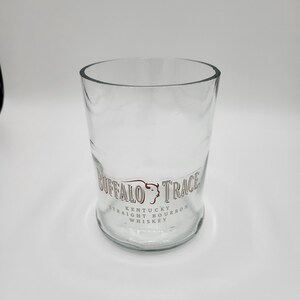 May include: A clear, cylindrical glass with the words "Buffalo Trace Kentucky Straight Bourbon Whiskey" printed in brown. The glass has a wide opening and a slightly curved base. The glass is empty.
