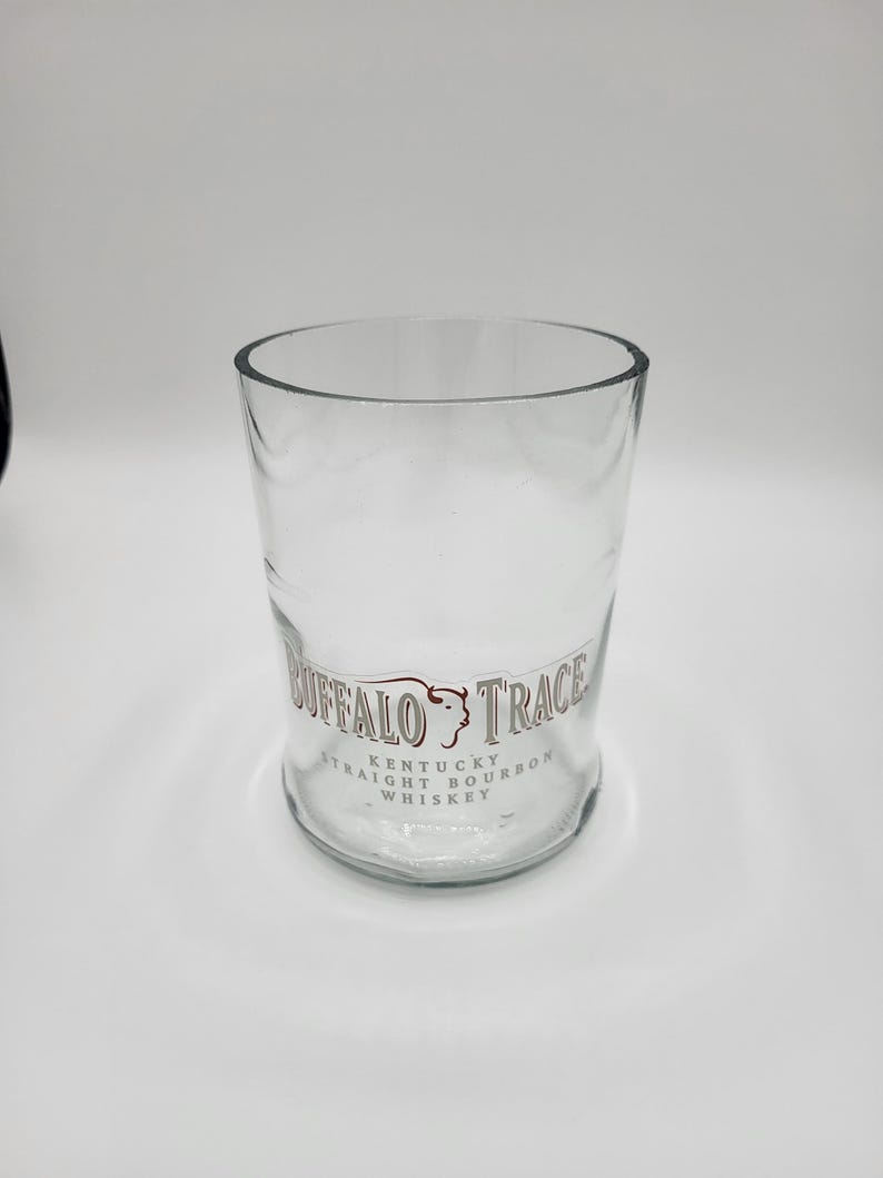 May include: A clear, cylindrical glass with the words "Buffalo Trace Kentucky Straight Bourbon Whiskey" printed in brown. The glass has a wide opening and a slightly curved base. The glass is empty.