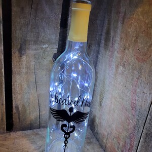 May include: A clear glass bottle with a cork top and white fairy lights inside. The bottle has a black decal with the text "Dedicated Nurse" and a medical symbol with wings.