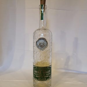 May include: A clear glass bottle with a green label that reads "Smoke Wagon Bottled in Bond Straight Rye Whiskey Las Vegas". The bottle has a textured surface and a cork stopper.