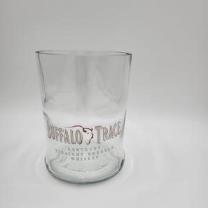 May include: A clear, cylindrical glass with a slightly curved body. The glass features the text "BUFFALO TRACE KENTUCKY STRAIGHT BOURBON WHISKEY" in a brown and white design. The glass is empty and sits on a white surface.