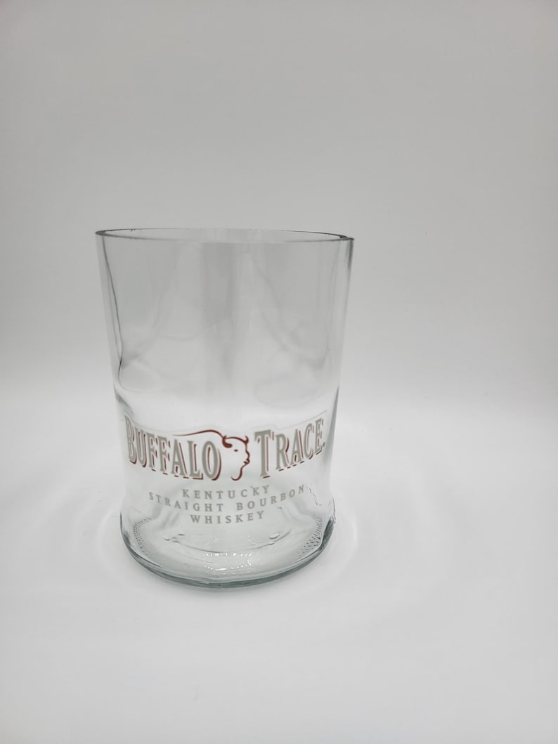 May include: A clear, cylindrical glass with a slightly curved body. The glass features the text "BUFFALO TRACE KENTUCKY STRAIGHT BOURBON WHISKEY" in a brown and white design. The glass is empty and sits on a white surface.