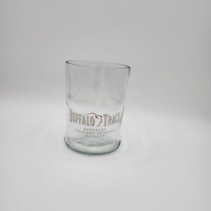 May include: A clear glass tumbler with a slanted top. The glass features the "Buffalo Trace Kentucky Straight Bourbon Whiskey" logo in brown lettering. The glass is designed for serving whiskey or other beverages.