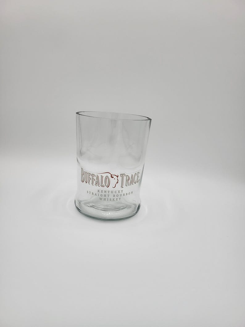 May include: A clear glass tumbler with a slanted top. The glass features the "Buffalo Trace Kentucky Straight Bourbon Whiskey" logo in brown lettering. The glass is designed for serving whiskey or other beverages.