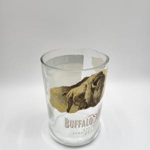 May include: Clear glass tumbler with a Buffalo Trace Kentucky Straight Whiskey label. The label features a brown illustration of a buffalo and the brand name in a serif font. The glass is cylindrical and appears to be repurposed.