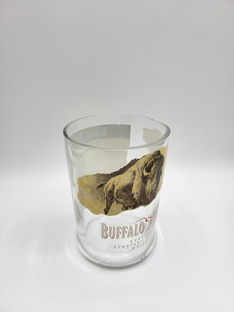 May include: Clear glass tumbler with a Buffalo Trace Kentucky Straight Whiskey label. The label features a brown illustration of a buffalo and the brand name in a serif font. The glass is cylindrical and appears to be repurposed.