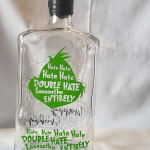 May include: Clear glass bottle with a black cap and a green graphic that says "Hate Hate, Double Hate, Looooathe Entirely". The bottle is decorated with string lights.