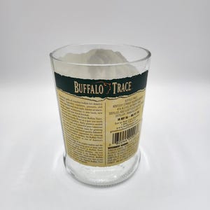 May include: A clear glass with the words "BUFFALO TRACE" in white and gold lettering. The glass has a dark green band at the top and is filled with a white substance. The glass has a label with text.