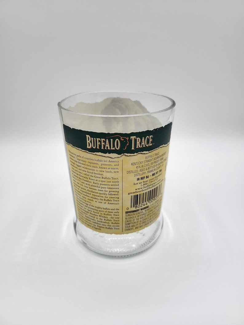 May include: A clear glass with the words "BUFFALO TRACE" in white and gold lettering. The glass has a dark green band at the top and is filled with a white substance. The glass has a label with text.