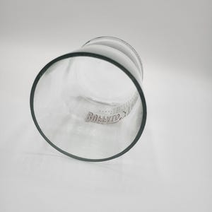 May include: A clear glass tumbler with a thick, dark rim. The glass has a cylindrical shape and is slightly tilted, revealing the interior. The word "Bollivio" is printed on the side of the glass.