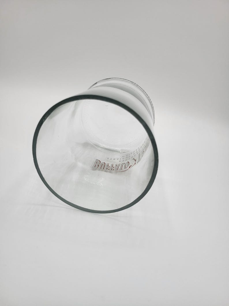 May include: A clear glass tumbler with a thick, dark rim. The glass has a cylindrical shape and is slightly tilted, revealing the interior. The word "Bollivio" is printed on the side of the glass.