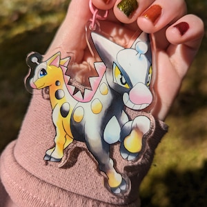 May include: A clear acrylic keyring featuring a colourful cartoon illustration of two creatures. One creature is yellow and black, the other is grey and white. The keyring has a pink clasp.
