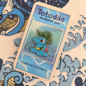 Puede incluir: Un pin de esmalte azul con el personaje Totodile, con una hoja verde y detalles rojos. El pin está en una tarjeta que dice "Totodile enamel pin" y "Pumpkin Plushness."