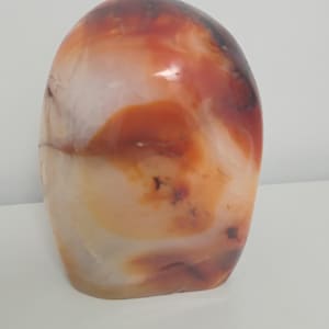 Peut inclure: Une pierre polie de forme ovale, présentant un mélange de couleurs. La pierre affiche des teintes blanches translucides, orangées et rouge foncé, créant un effet marbré. La surface lisse reflète la lumière, soulignant les motifs naturels.
