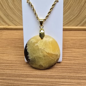 Pode incluir: Um colar de corrente dourada com um pendente grande e redondo. O pendente apresenta tons amarelos e castanhos, com uma superfície texturizada. O colar é exibido em um suporte branco, sobre um fundo de madeira.