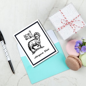 May include: A white card with a blue border and a black and white illustration of a lamb with a flag and the text "Agnus Dei". The card is sitting on a light blue envelope.