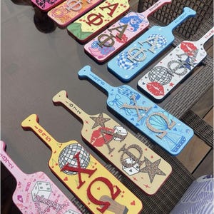 May include: A collection of ten decorative wooden paddles arranged in rows, each featuring unique hand-painted designs, Greek letters embellished with rhinestones or patterns, and various motifs including disco balls, dice, lips, and cowboy boots.