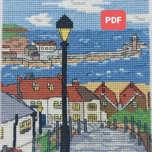 May include: A cross-stitch pattern of a seaside town with a lamppost in the foreground and a lighthouse in the background. The pattern is titled "Whitby" and is available as a PDF download.