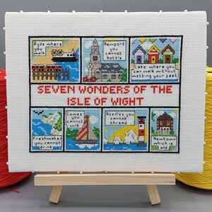 May include: A white cross-stitch embroidery on a wooden easel depicting the Seven Wonders of the Isle of Wight. The embroidery is divided into seven panels, each with a different illustration and a short description. The panels depict Ryde, Newport, Freshwater, Cowes, Needles, Lake, and Newtown.