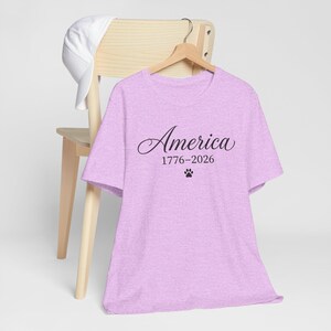 Könnte beinhalten: Ein helllila T-Shirt mit dem Wort "America" in Schreibschrift, den Daten "1776-2026" und einem Pfotenabdruck. Eine weiße Baseballkappe liegt auf einem Holzstuhl hinter dem Shirt.