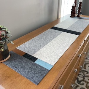May include: A gray, blue, and brown felt table runner with a geometric pattern. The runner is on a wooden table with drawers.