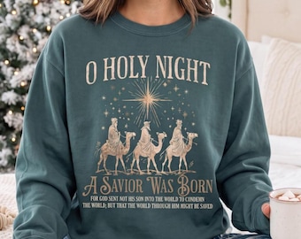 May include: A dark teal crewneck sweatshirt featuring a graphic print of three figures riding camels under a star. The text reads O HOLY NIGHT, A SAVIOR WAS BORN, and FOR GOD SENT NOT HIS SON INTO THE WORLD TO CONDEMN THE WORLD; BUT THAT THE WORLD THROUGH HIM MIGHT BE SAVED.