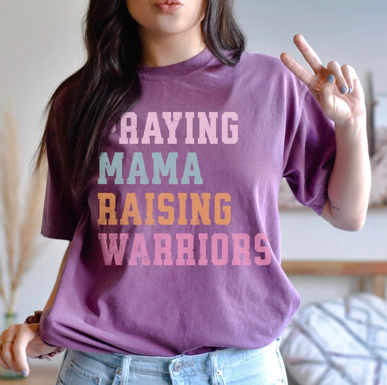 May include: A purple t-shirt with the text 'Praying Mama Raising Warriors' in a stacked vertical design. The text is in different colors, including pink, orange, blue, and white. Praying Mama, Comfort Colors Tshirt, Christian Shirts, Mama Shirt, Mom Shirt, Mothers Day Gift, Gifts For Mom, Christian Apparel, Gift For Wife, Religious Shirt, Religious Gifts for Her, New Mom Gift. Mom Gifts, Christian Gifts