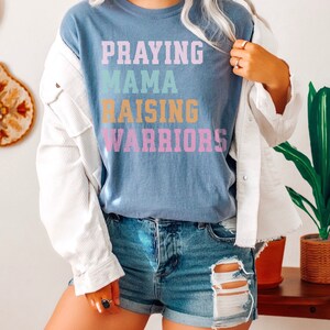 May include: A blue t-shirt with the text 'Praying Mama Raising Warriors' in rainbow colors. Praying Mama, Comfort Colors Tshirt, Christian Shirts, Mama Shirt, Mom Shirt, Mothers Day Gift, Gifts For Mom, Christian Apparel, Gift For Wife, Religious Shirt, Religious Gifts for Her, New Mom Gift. Mom Gifts, Christian Gifts