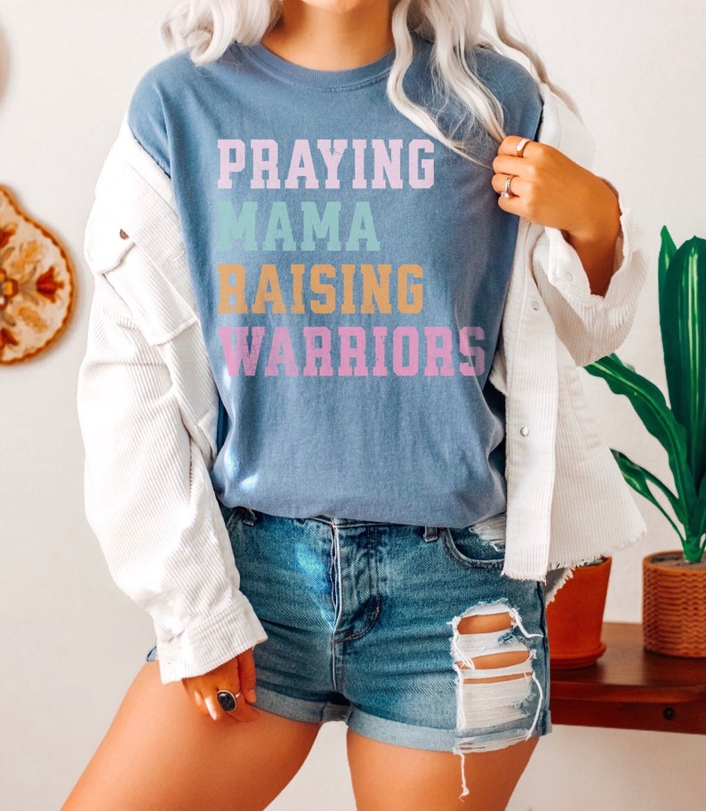 May include: A blue t-shirt with the text 'Praying Mama Raising Warriors' in rainbow colors. Praying Mama, Comfort Colors Tshirt, Christian Shirts, Mama Shirt, Mom Shirt, Mothers Day Gift, Gifts For Mom, Christian Apparel, Gift For Wife, Religious Shirt, Religious Gifts for Her, New Mom Gift. Mom Gifts, Christian Gifts