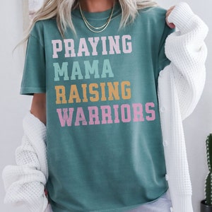 May include: A teal blue t-shirt with the text 'Praying Mama Raising Warriors' in a retro font. The text is in different colors: pink, yellow, and light blue. Praying Mama, Comfort Colors Tshirt, Christian Shirts, Mama Shirt, Mom Shirt, Mothers Day Gift, Gifts For Mom, Christian Apparel, Gift For Wife, Religious Shirt, Religious Gifts for Her, New Mom Gift. Mom Gifts, Christian Gifts