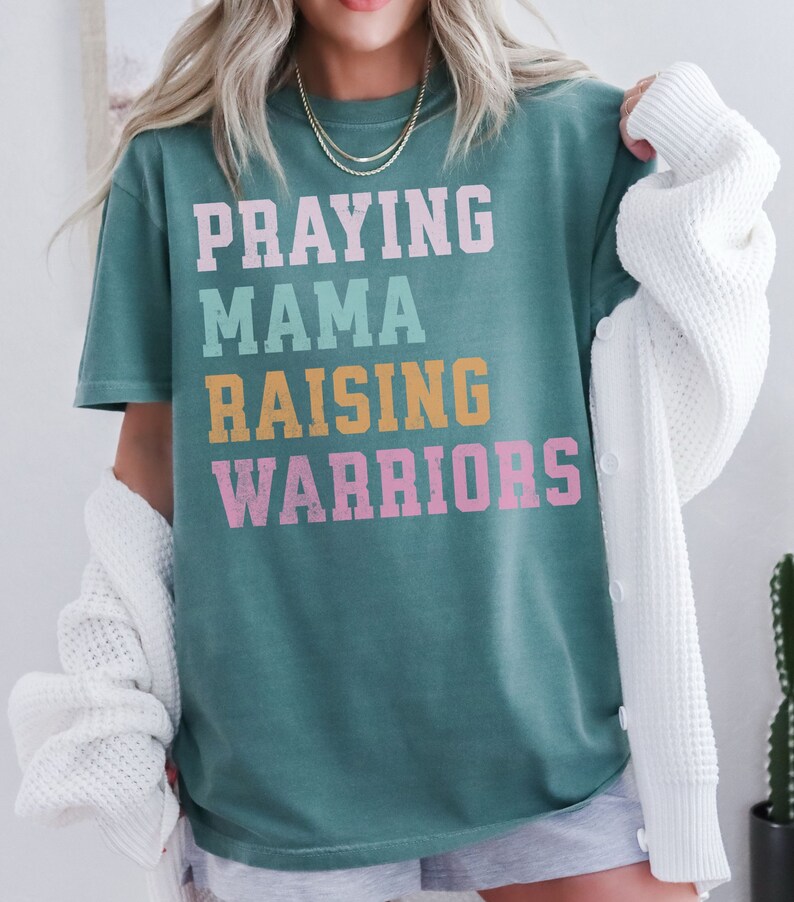 May include: A teal blue t-shirt with the text 'Praying Mama Raising Warriors' in a retro font. The text is in different colors: pink, yellow, and light blue. Praying Mama, Comfort Colors Tshirt, Christian Shirts, Mama Shirt, Mom Shirt, Mothers Day Gift, Gifts For Mom, Christian Apparel, Gift For Wife, Religious Shirt, Religious Gifts for Her, New Mom Gift. Mom Gifts, Christian Gifts