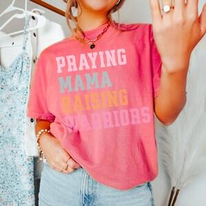 May include: A pink t-shirt with the text 'Praying Mama Raising Warriors' in colorful letters. Praying Mama, Comfort Colors Tshirt, Christian Shirts, Mama Shirt, Mom Shirt, Mothers Day Gift, Gifts For Mom, Christian Apparel, Gift For Wife, Religious Shirt, Religious Gifts for Her, New Mom Gift. Mom Gifts, Christian Gifts