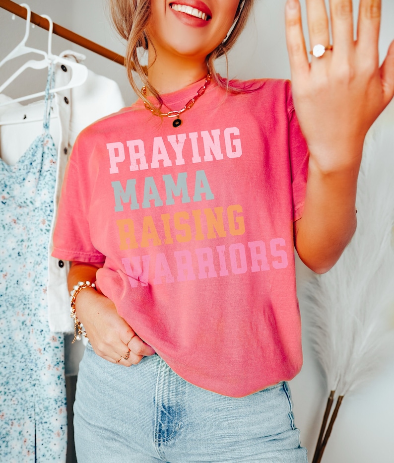 May include: A pink t-shirt with the text 'Praying Mama Raising Warriors' in colorful letters. Praying Mama, Comfort Colors Tshirt, Christian Shirts, Mama Shirt, Mom Shirt, Mothers Day Gift, Gifts For Mom, Christian Apparel, Gift For Wife, Religious Shirt, Religious Gifts for Her, New Mom Gift. Mom Gifts, Christian Gifts