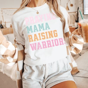 May include: A white t-shirt with the text 'Praying Mama Raising Warriors' in colorful letters. The letters are in pink, blue, orange, and pink. The shirt is being worn by a person. Praying Mama, Comfort Colors Tshirt, Christian Shirts, Mama Shirt, Mom Shirt, Mothers Day Gift, Gifts For Mom, Christian Apparel, Gift For Wife, Religious Shirt, Religious Gifts for Her, New Mom Gift. Mom Gifts, Christian Gifts