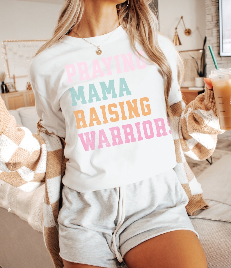 May include: A white t-shirt with the text 'Praying Mama Raising Warriors' in colorful letters. The letters are in pink, blue, orange, and pink. The shirt is being worn by a person. Praying Mama, Comfort Colors Tshirt, Christian Shirts, Mama Shirt, Mom Shirt, Mothers Day Gift, Gifts For Mom, Christian Apparel, Gift For Wife, Religious Shirt, Religious Gifts for Her, New Mom Gift. Mom Gifts, Christian Gifts