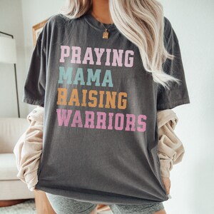 May include: A gray t-shirt with the text 'Praying Mama Raising Warriors' in colorful letters. The letters are in pink, blue, orange, and white. Praying Mama, Comfort Colors Tshirt, Christian Shirts, Mama Shirt, Mom Shirt, Mothers Day Gift, Gifts For Mom, Christian Apparel, Gift For Wife, Religious Shirt, Religious Gifts for Her, New Mom Gift. Mom Gifts, Christian Gifts