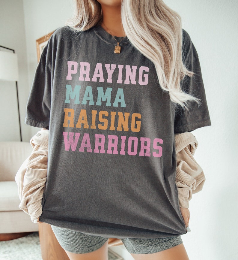 May include: A gray t-shirt with the text 'Praying Mama Raising Warriors' in colorful letters. The letters are in pink, blue, orange, and white. Praying Mama, Comfort Colors Tshirt, Christian Shirts, Mama Shirt, Mom Shirt, Mothers Day Gift, Gifts For Mom, Christian Apparel, Gift For Wife, Religious Shirt, Religious Gifts for Her, New Mom Gift. Mom Gifts, Christian Gifts