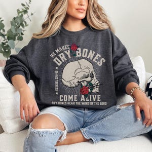 May include: A dark gray sweatshirt with a white skull and crossbones graphic. The graphic includes the text "He Makes Dry Bones Come Alive" and a Bible verse reference "Ezekiel 37:1-10".