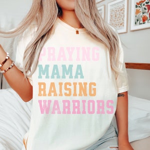 May include: A white t-shirt with the text 'Praying Mama Raising Warriors' in pink, blue, and orange lettering. Praying Mama, Comfort Colors Tshirt, Christian Shirts, Mama Shirt, Mom Shirt, Mothers Day Gift, Gifts For Mom, Christian Apparel, Gift For Wife, Religious Shirt, Religious Gifts for Her, New Mom Gift. Mom Gifts, Christian Gifts